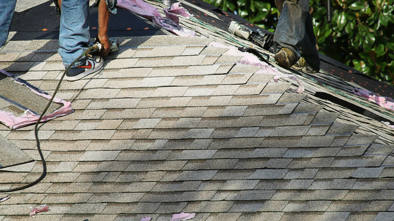 Roofer using a nail gun on light brown shingles near the ridge